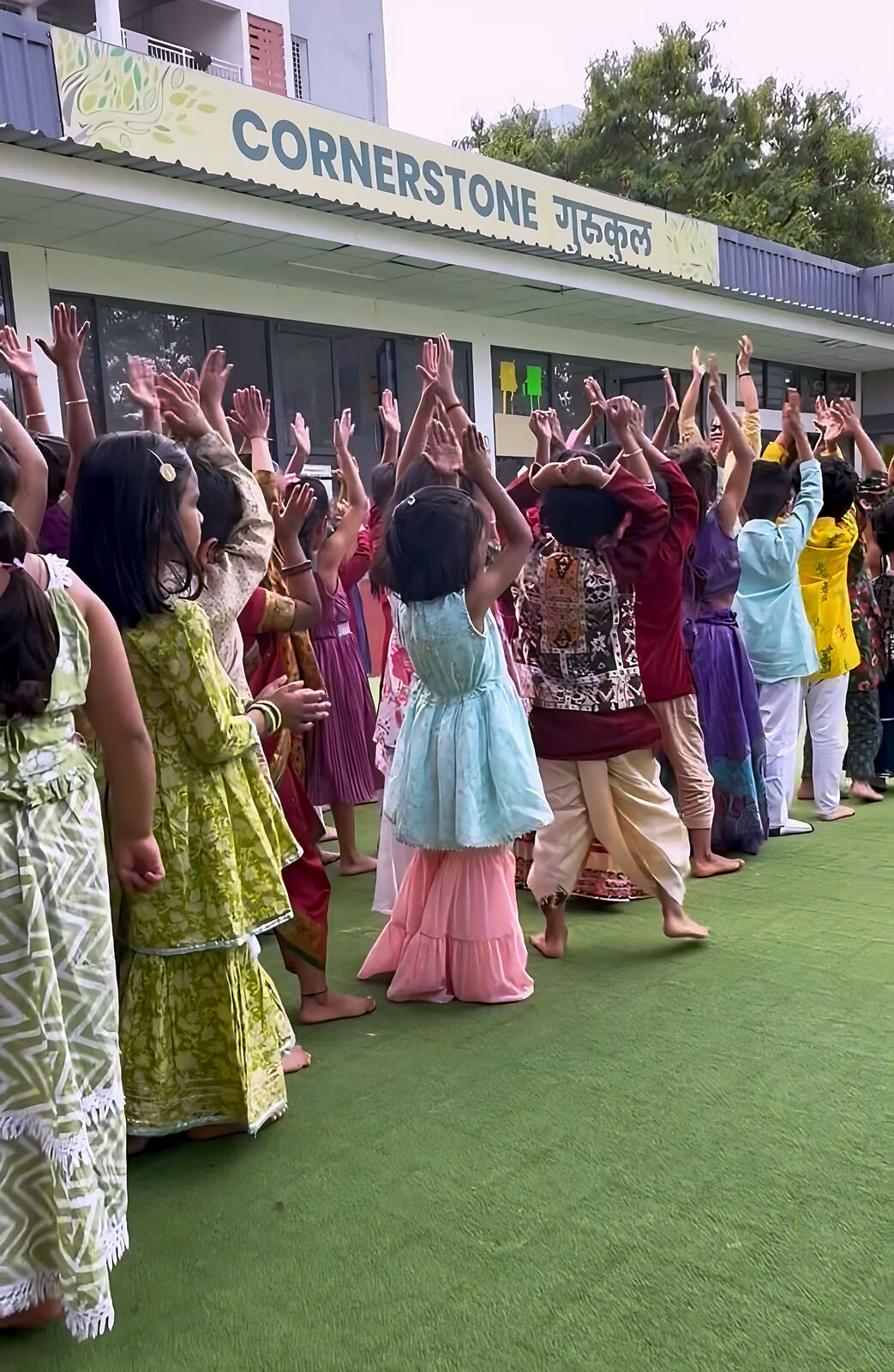 Children and teachers raise their arms on the green turf in front of the Cornerstone Gurukul building