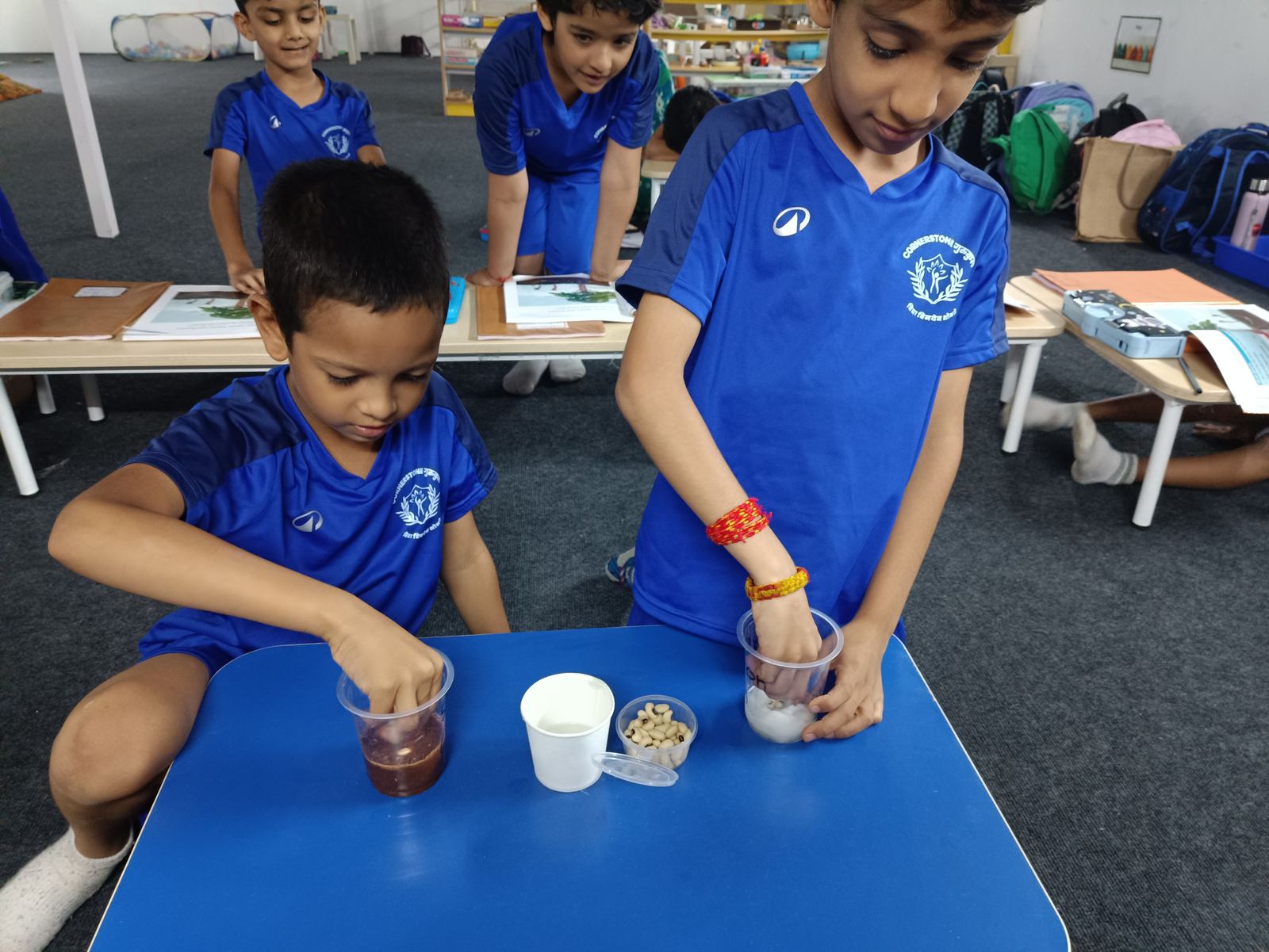 Two children in blue kit pour liquid into small cups at a blue table, watched by classmates