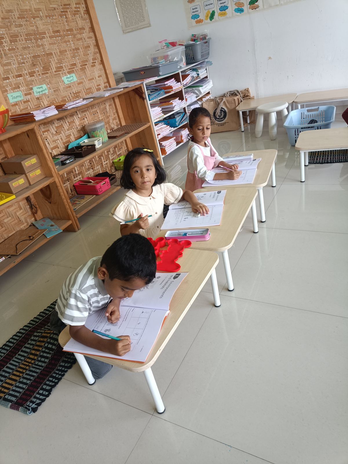Three children write at a long low desk, each on their own sheet, in a bright classroom