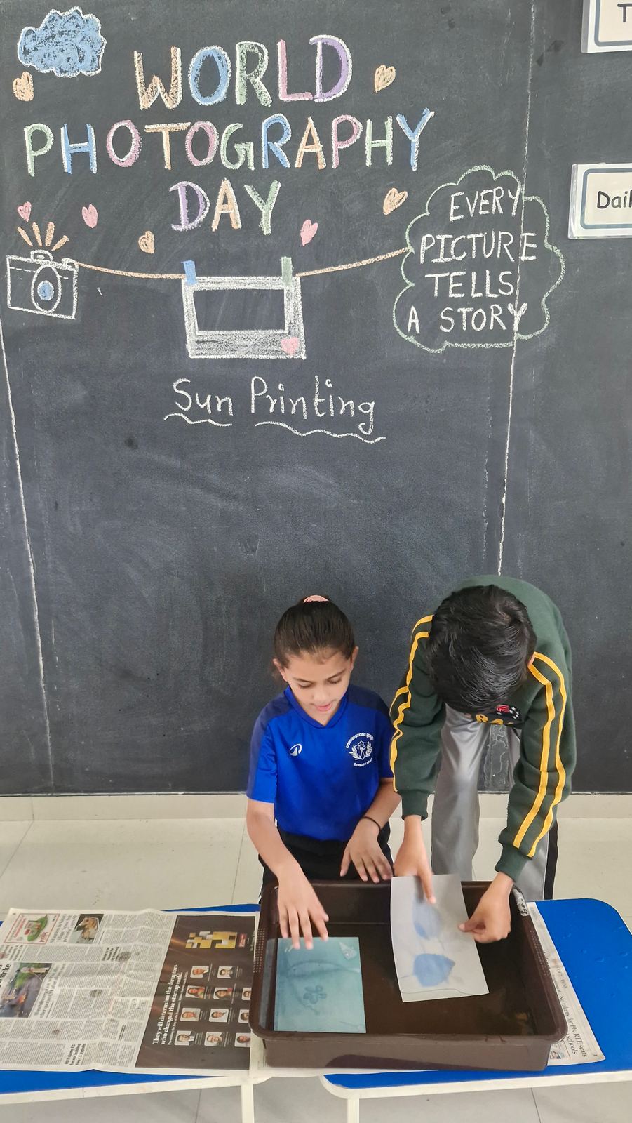 Two children in blue kit make a sun print together over a tray, one guiding the other’s hands