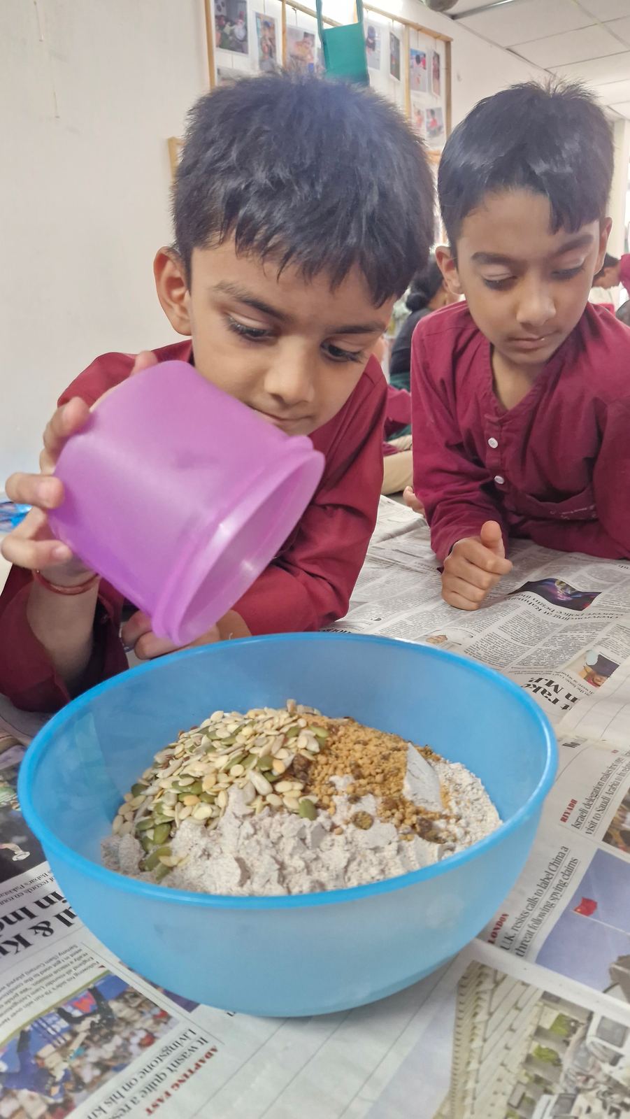Two boys in maroon kurtas pour seeds into a bowl of laddoo mix together