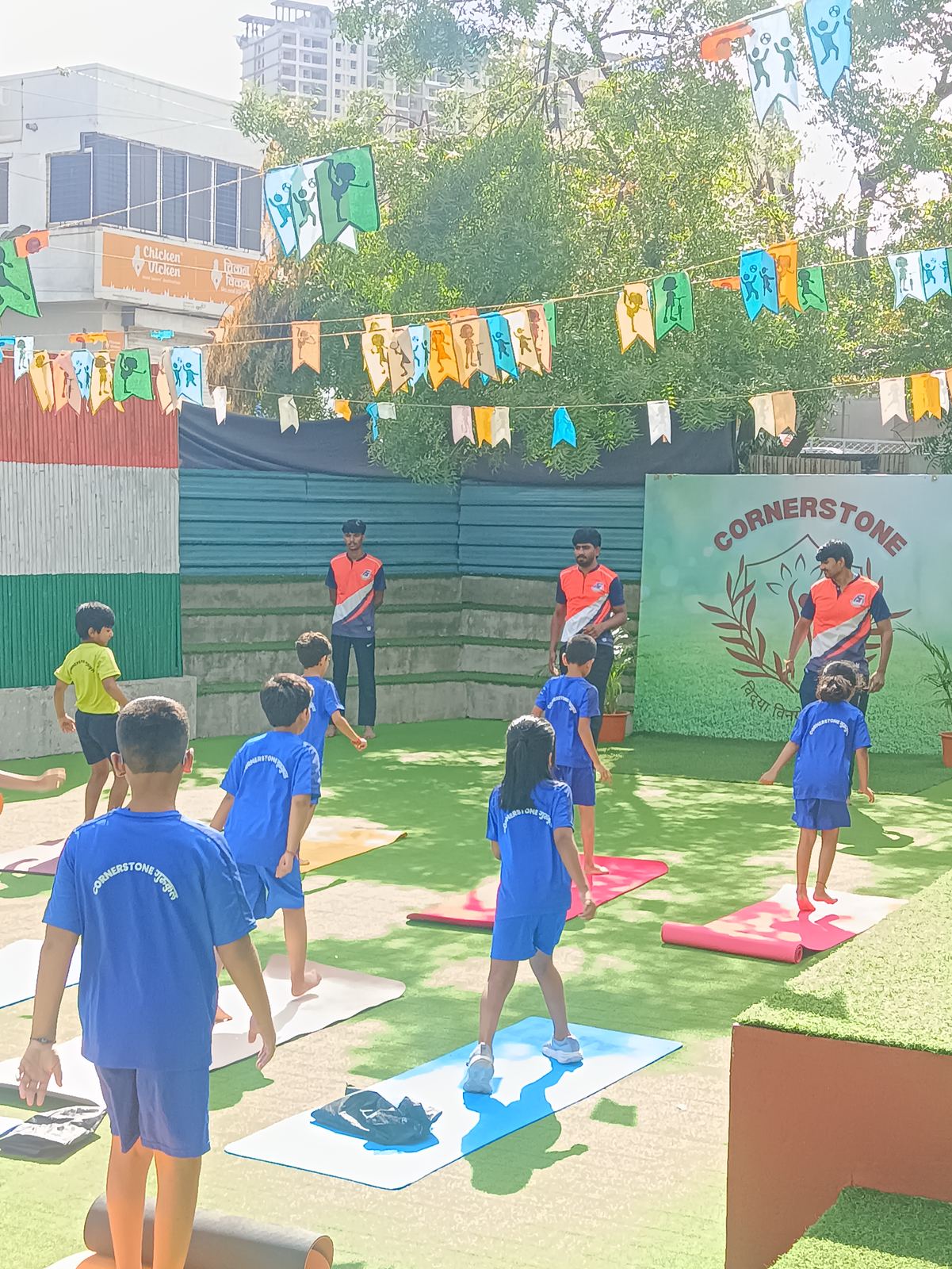 Children in blue sports kit stretch their arms overhead on yoga mats on the turf, under bunting